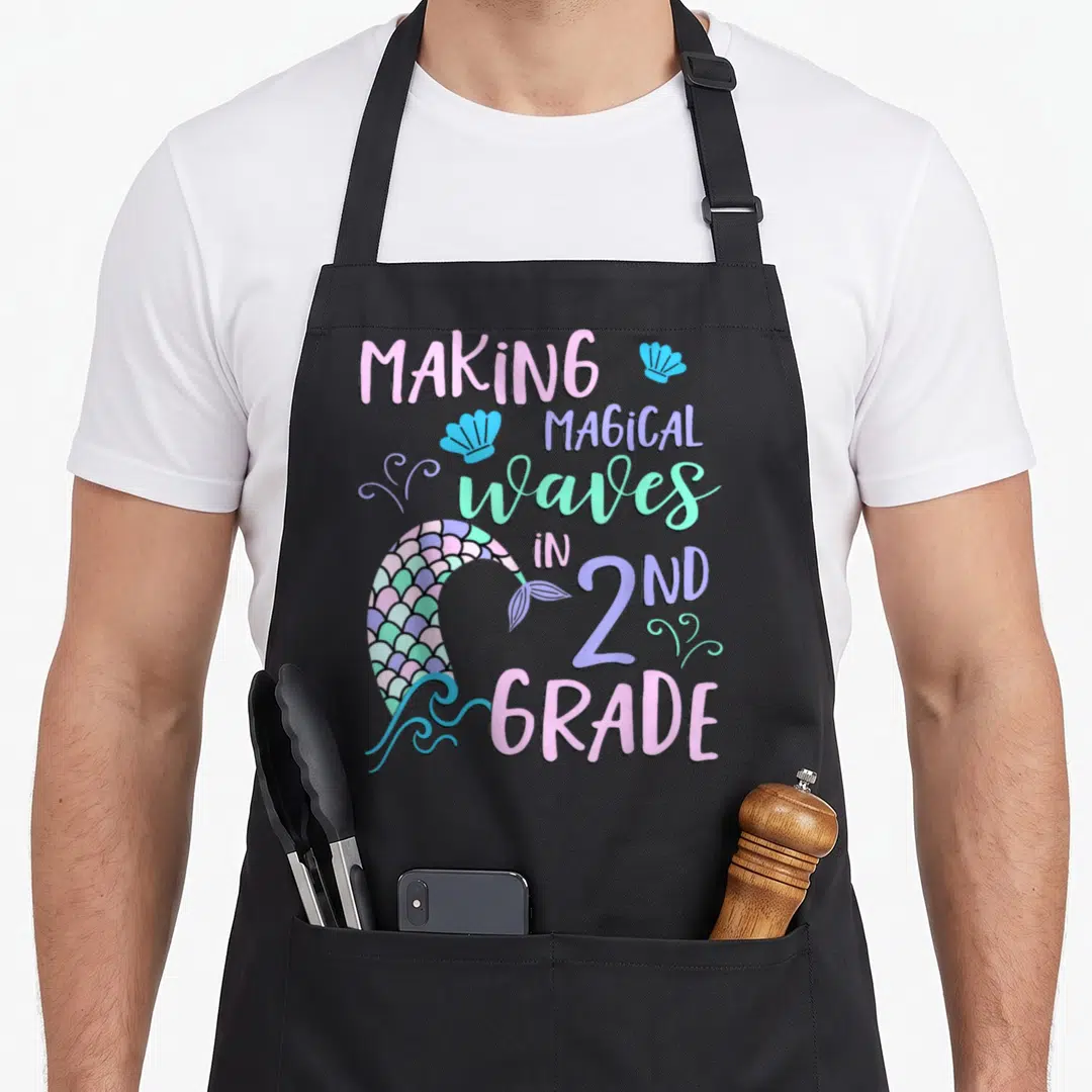 Grade 2nd apron - Making Magical Waves in 2nd Grade text with mermaid tail and seashells — shown on a black cotton canvas apron against a white background