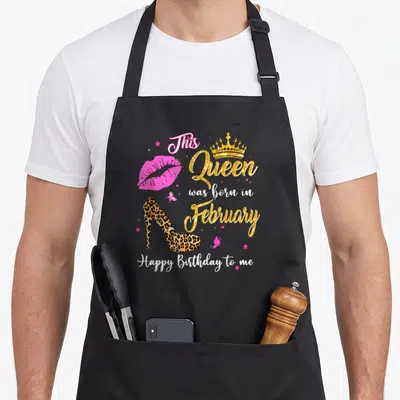 February leopard apron - Gold crown, pink lips, leopard heel, stars, butterflies, and text 'This Queen was born — shown on a black cotton canvas apron against a white background