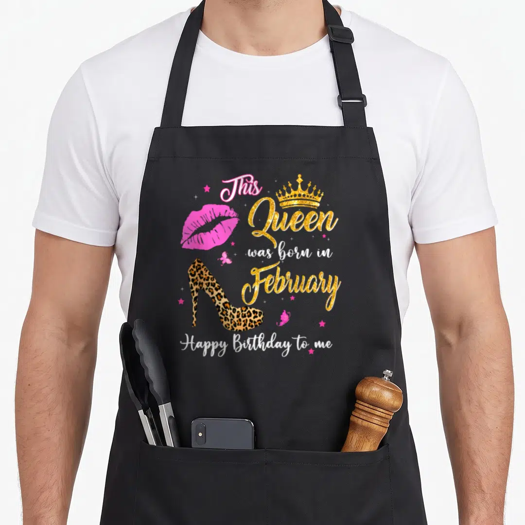 February leopard apron - Gold crown, pink lips, leopard heel, stars, butterflies, and text 'This Queen was born — shown on a black cotton canvas apron against a white background