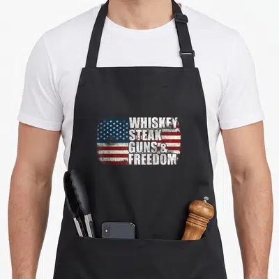 Whiskey 4th of july apron - Distressed USA flag with stacked text WHISKEY STEAK GUNS FREEDOM in red white blue — shown on a black cotton canvas apron against a white background