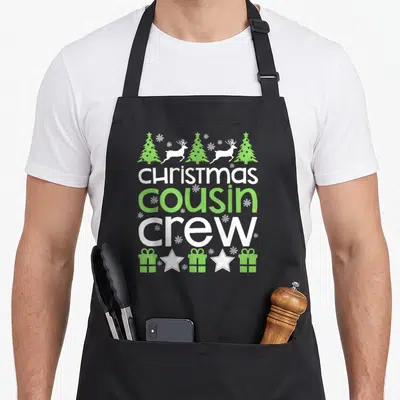 Crew family christmas aprons - Stacked text Christmas cousin in crew with reindeer trees and gifts in white and lime — shown on a black cotton canvas apron against a white background
