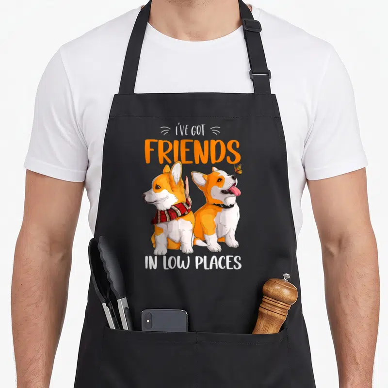 Friend pembroke corgi aprons - Two corgis sitting with scarf and butterfly under friends in low places text — shown on a black cotton canvas apron against a white background