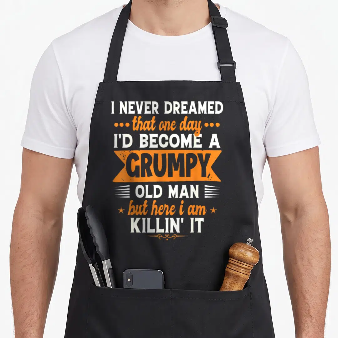 Dreamed father humor apron - Stacked white and orange typography reading I Never Dreamed I'd Become A Grumpy Old Man — shown on a black cotton canvas apron against a white background