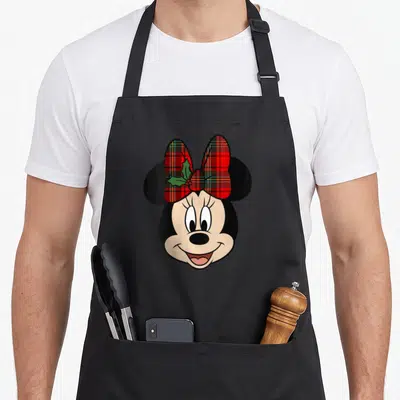 Mistletoe christmas apron - Cartoon Minnie Mouse head with red plaid bow and green holly leaves — shown on a black cotton canvas apron against a white background