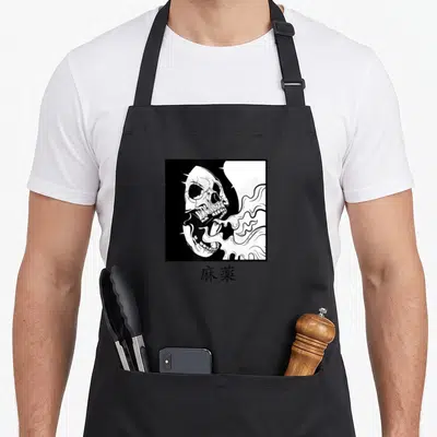 Japanese minimalist apron - Black and white skull smoking inside square frame with kanji text below — shown on a black cotton canvas apron against a white background