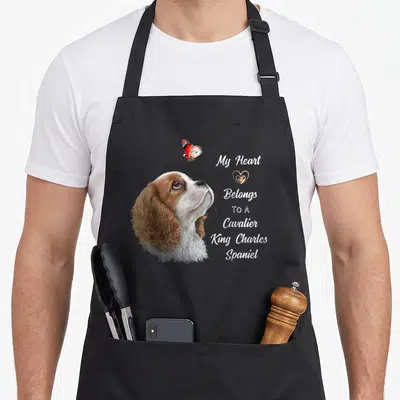 Spaniel dog king apron - Realistic brown and white dog head looking up at orange butterfly beside stacked white text — shown on a black cotton canvas apron against a white background