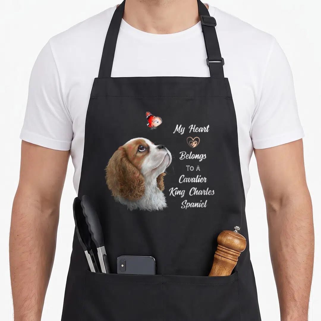 Spaniel dog king apron - Realistic brown and white dog head looking up at orange butterfly beside stacked white text — shown on a black cotton canvas apron against a white background