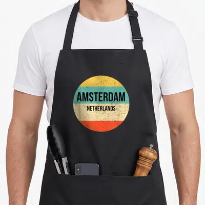 Retro amsterdam netherlands apron - Circular graphic with yellow teal beige and red bands containing stacked black — shown on a black cotton canvas apron against a white background