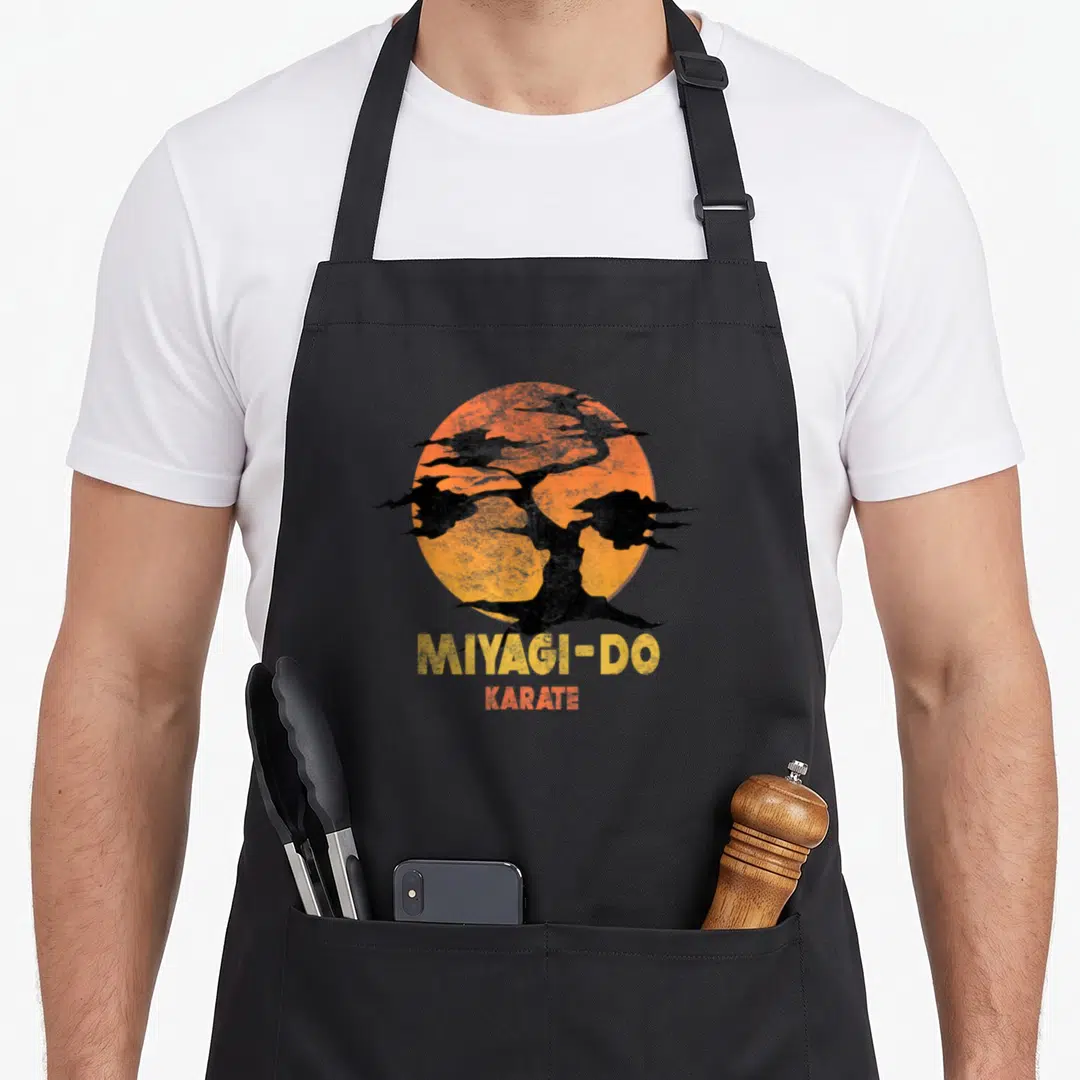 Karate tree apron - Black bonsai silhouette on orange sun with stacked MIYAGI-DO KARATE text in yellow and red — shown on a black cotton canvas apron against a white background