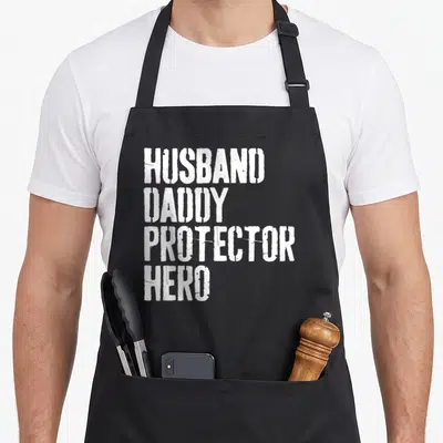 Hero daddy aprons - White stacked text reading HUSBAND DADDY PROTECTOR HERO on black fabric — shown on a black cotton canvas apron against a white background