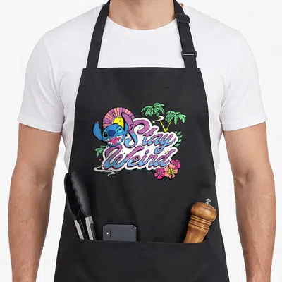Stay weird aprons - Blue cartoon character winking beside Stay Weird script text with green palms and pink flowers — shown on a black cotton canvas apron against a white background
