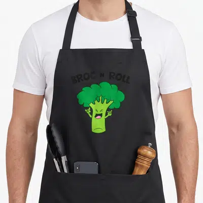 Roll vegetable apron - Green cartoon broccoli with raised arms under arched black text BROC N ROLL — shown on a black cotton canvas apron against a white background
