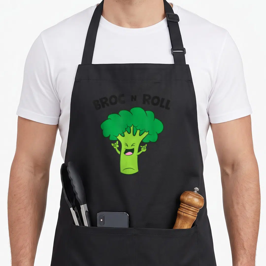 Roll vegetable apron - Green cartoon broccoli with raised arms under arched black text BROC N ROLL — shown on a black cotton canvas apron against a white background