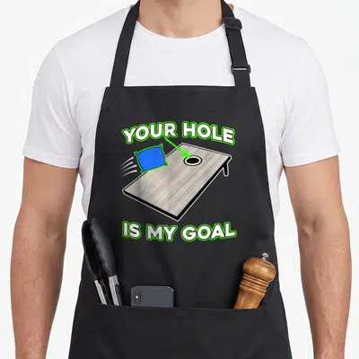 Cornhole yard apron - Cartoon brown cornhole board with blue bean bag, green arrow, and white text outlined in green — shown on a black cotton canvas apron against a white background