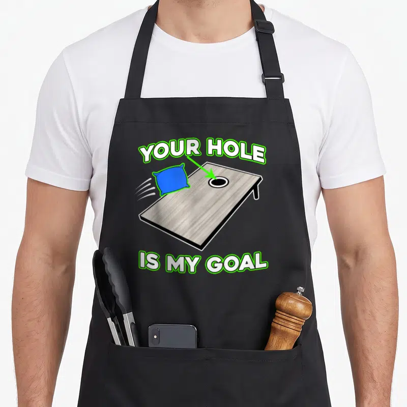 Cornhole yard apron - Cartoon brown cornhole board with blue bean bag, green arrow, and white text outlined in green — shown on a black cotton canvas apron against a white background
