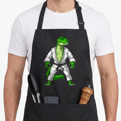 Jiu dinosaur apron - Green dinosaur standing in white gi and black belt martial arts cartoon — shown on a black cotton canvas apron against a white background