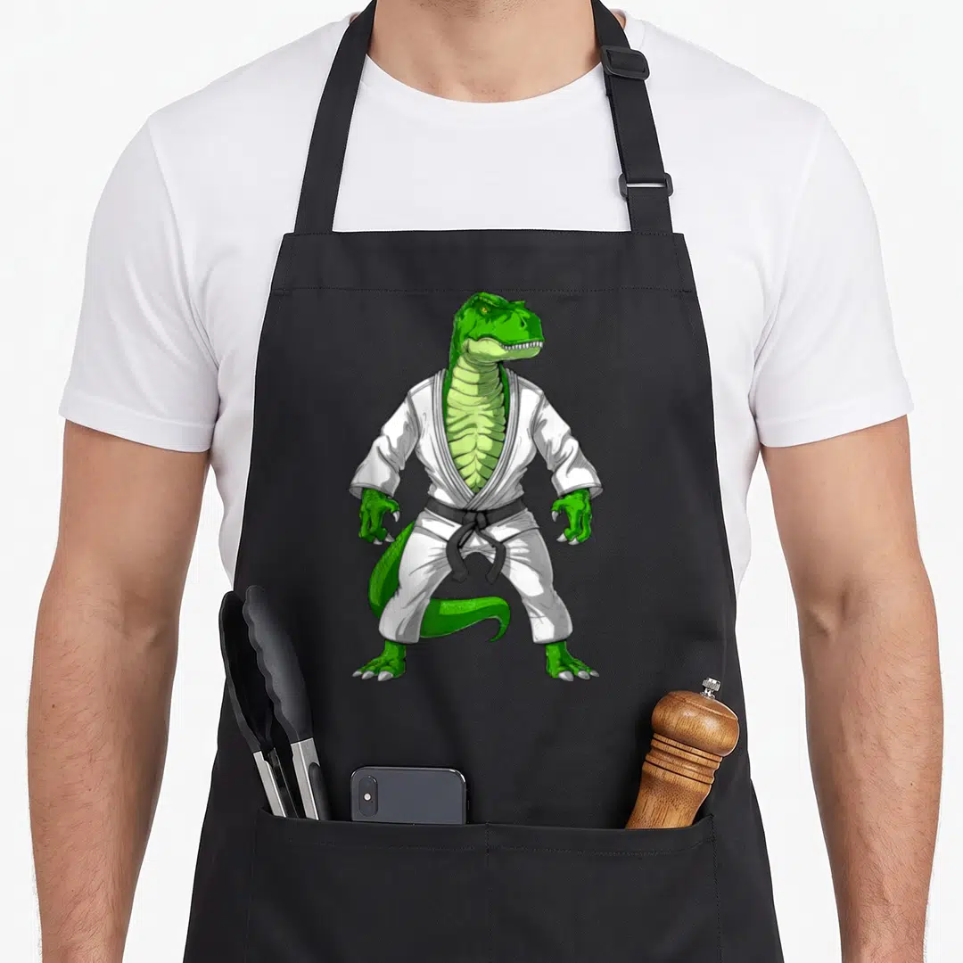 Jiu dinosaur apron - Green dinosaur standing in white gi and black belt martial arts cartoon — shown on a black cotton canvas apron against a white background
