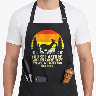 See bow apron - Distressed deer sunset silhouette with text listing deer jerky steaks sausages and burgers — shown on a black cotton canvas apron against a white background