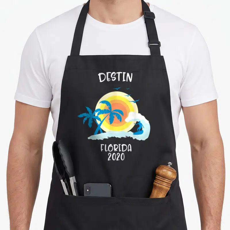 Destin family aprons - Cartoon beach scene with surfer, two palm trees, yellow sun, and white text reading Destin — shown on a black cotton canvas apron against a white background