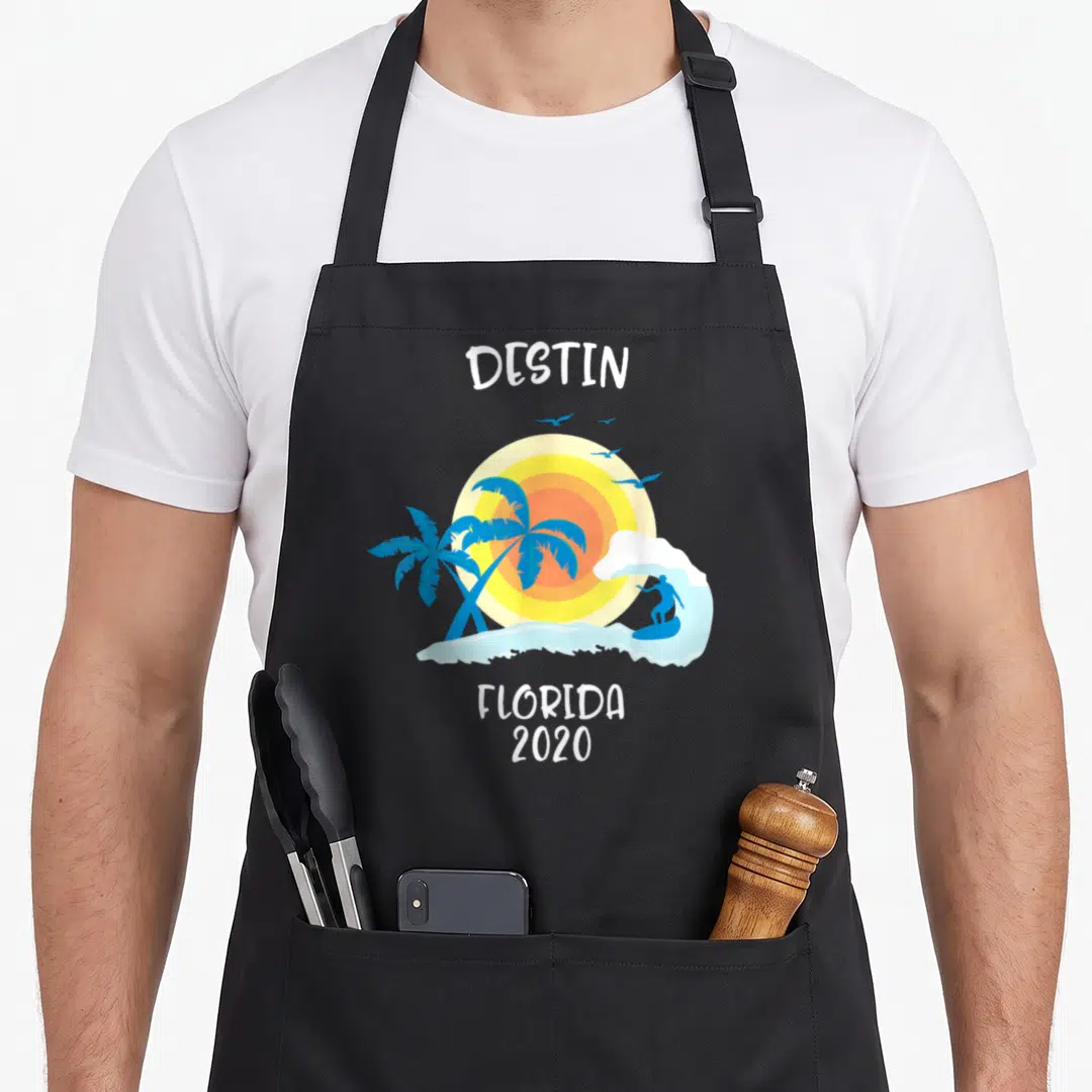 Destin family aprons - Cartoon beach scene with surfer, two palm trees, yellow sun, and white text reading Destin — shown on a black cotton canvas apron against a white background
