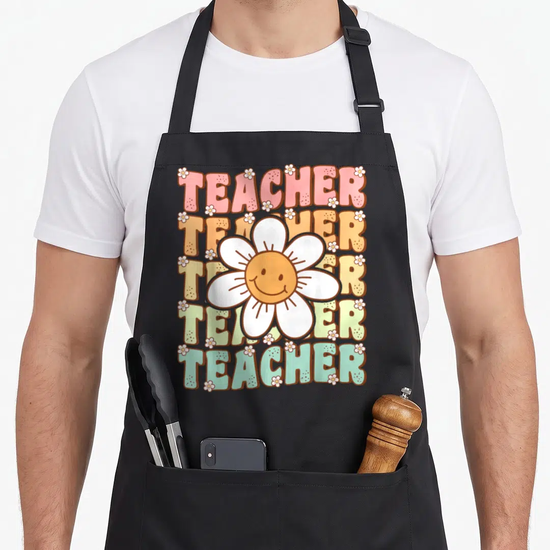 Daisy teacher apron - Five stacked TEACHER words in pink orange yellow green and white with a smiling white daisy — shown on a black cotton canvas apron against a white background