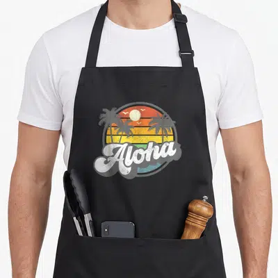 Vacation hawaiian apron - Retro sunset with Aloha text, palm trees, birds, orange and yellow stripes — shown on a black cotton canvas apron against a white background