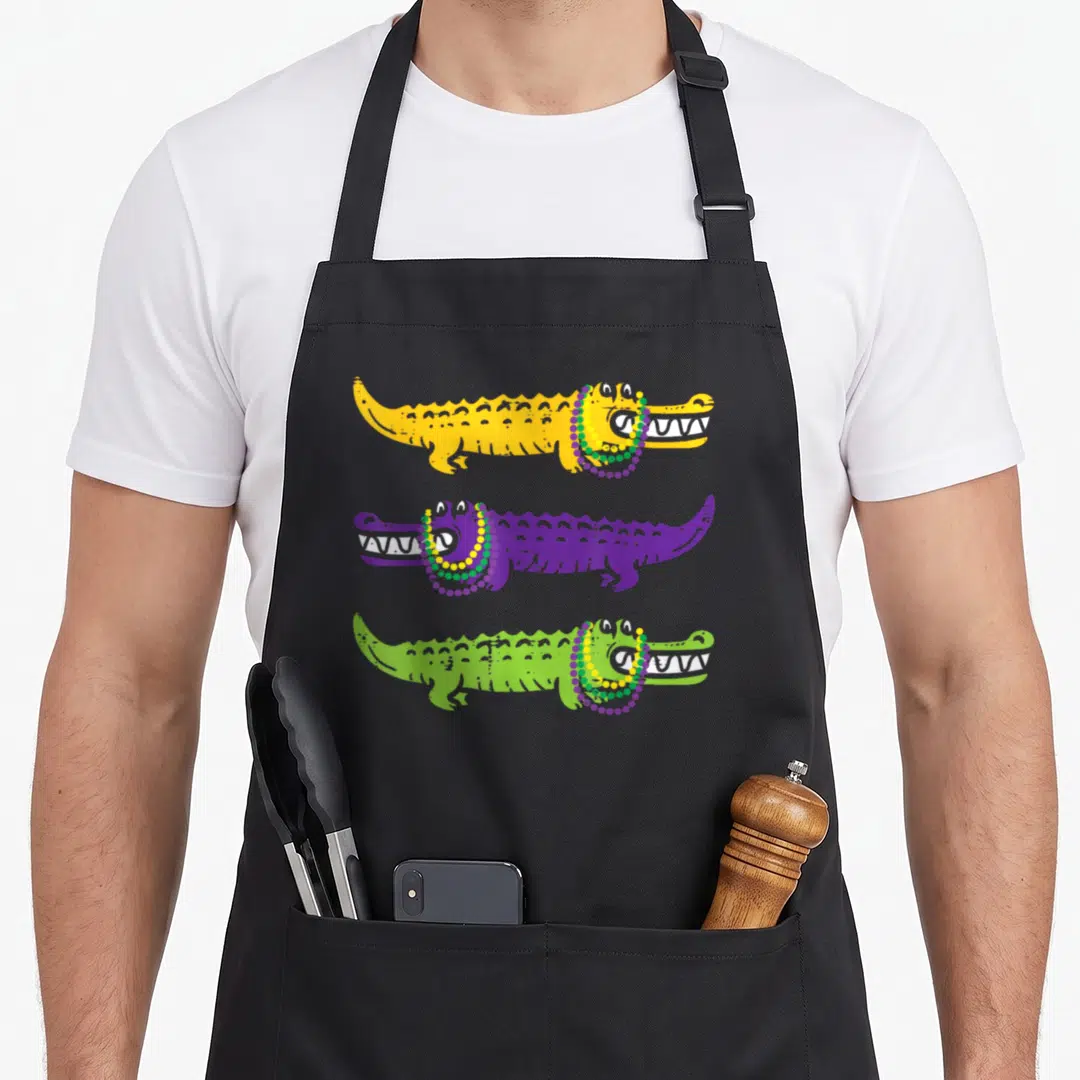 Aligator louisiana apron - Three cartoon alligators in yellow purple and green wearing Mardi Gras beads — shown on a black cotton canvas apron against a white background