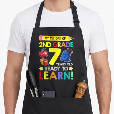Grade school 2nd apron - Pencil with text, number 7, apple, books, backpack with cap, colorful lettering — shown on a black cotton canvas apron against a white background