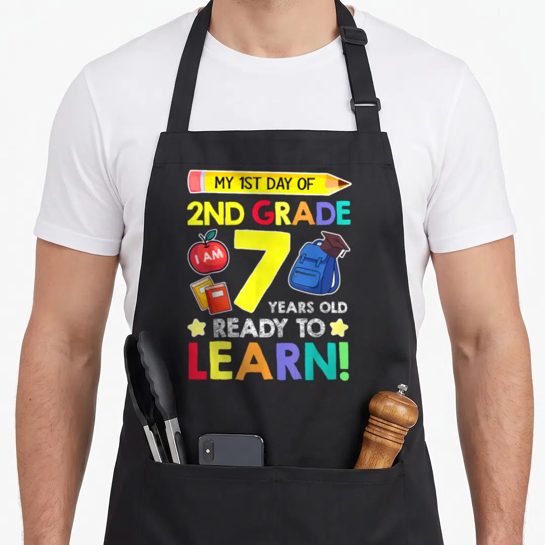 Grade school 2nd apron - Pencil with text, number 7, apple, books, backpack with cap, colorful lettering — shown on a black cotton canvas apron against a white background