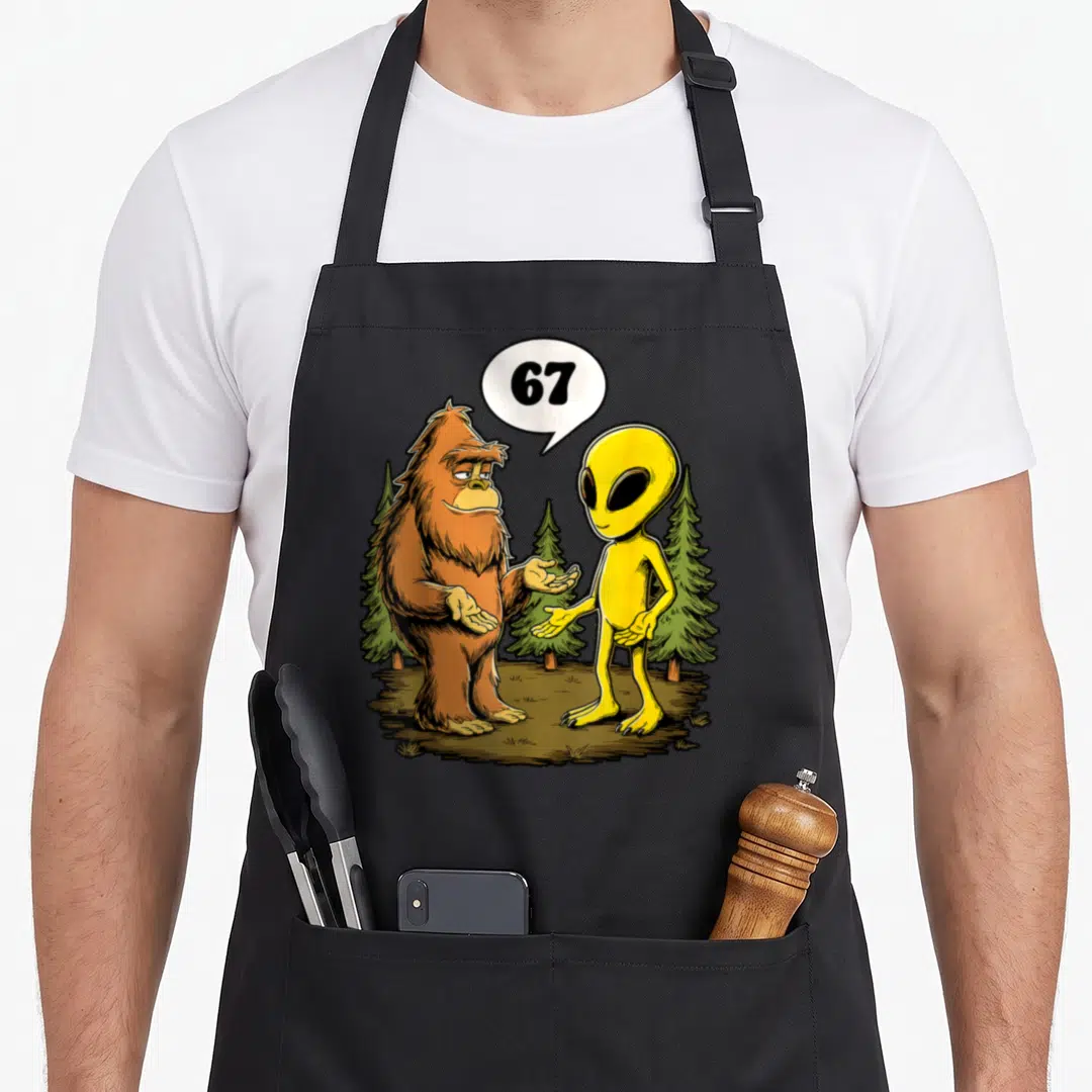 Six bigfoot apron - Bigfoot and yellow alien shaking hands under speech bubble with number 67 and pine trees — shown on a black cotton canvas apron against a white background