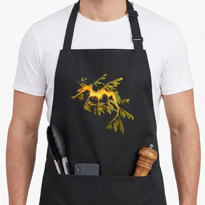 Leafy animal apron - Realistic yellow and green leafy seadragon illustration with leaf-shaped fins — shown on a black cotton canvas apron against a white background