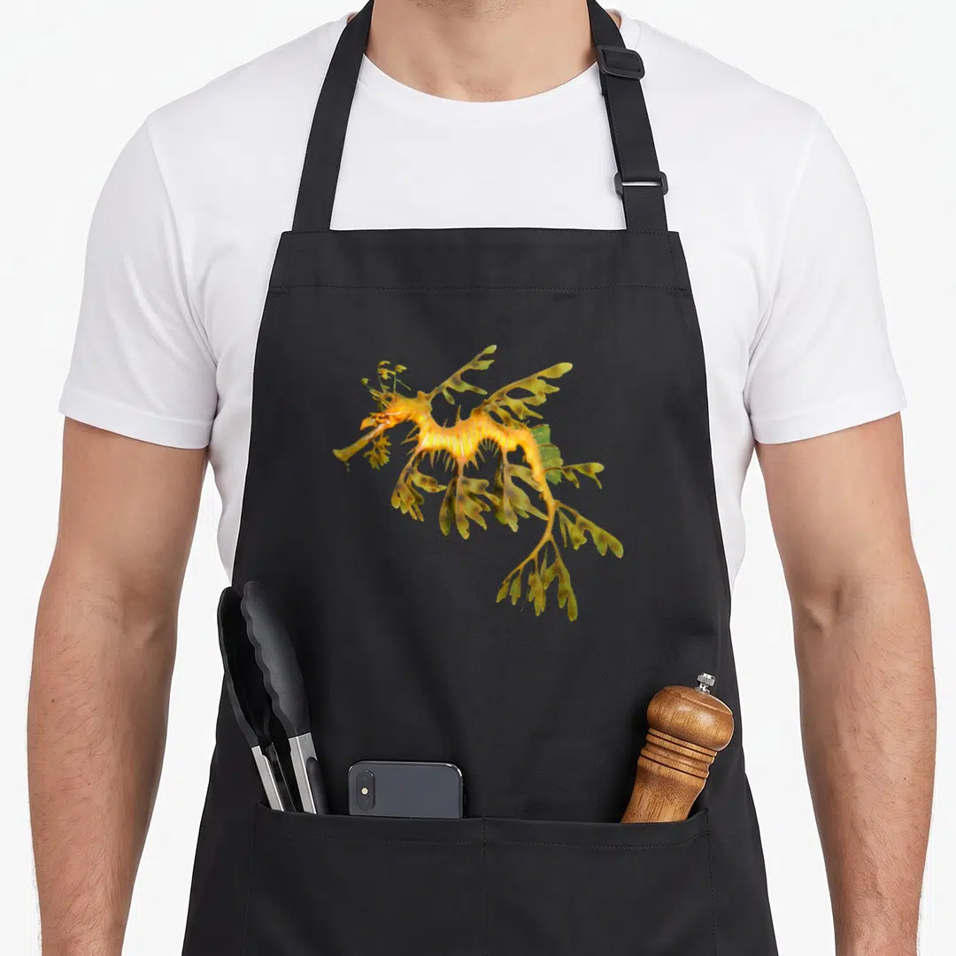 Leafy animal apron - Realistic yellow and green leafy seadragon illustration with leaf-shaped fins — shown on a black cotton canvas apron against a white background