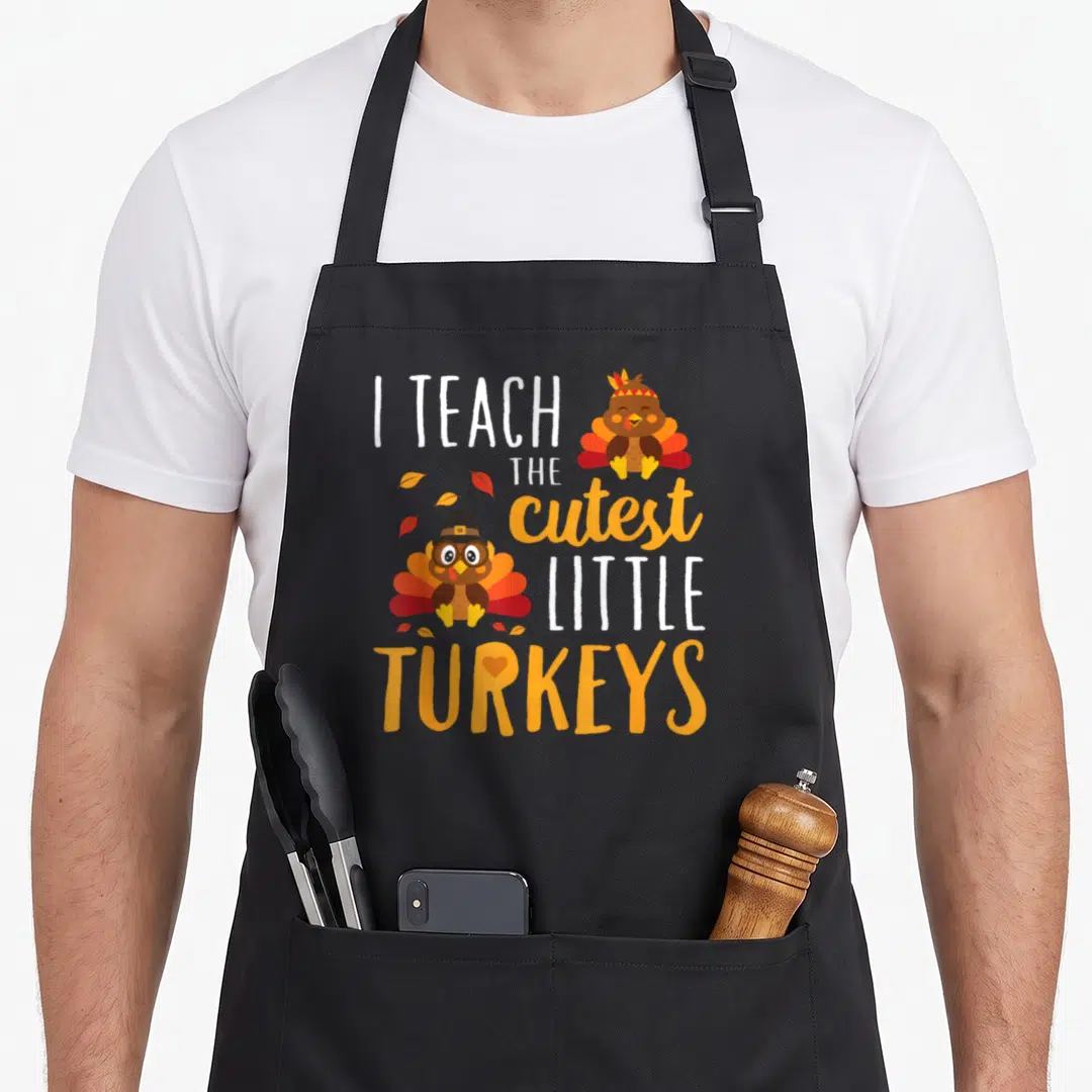 Teach thankful apron - I TEACH THE cutest LITTLE TURKEYS text with two cartoon turkeys and autumn leaves — shown on a black cotton canvas apron against a white background