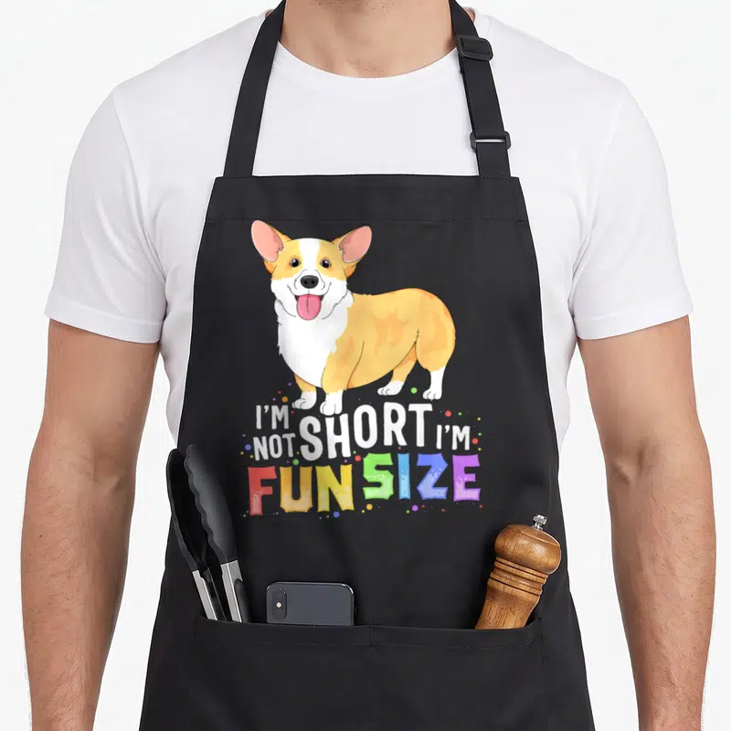 Funny dog corgi apron - Cartoon corgi with I'm Not Short I'm Fun Size text and colorful dots — shown on a black cotton canvas apron against a white background