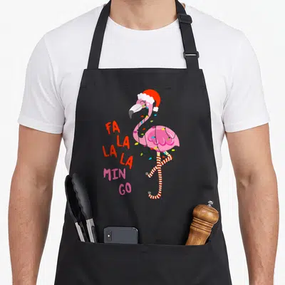 Flamingo christmas tree apron - Pink flamingo wearing red santa hat and wrapped in lights beside FA LA LA MIN GO text — shown on a black cotton canvas apron against a white background