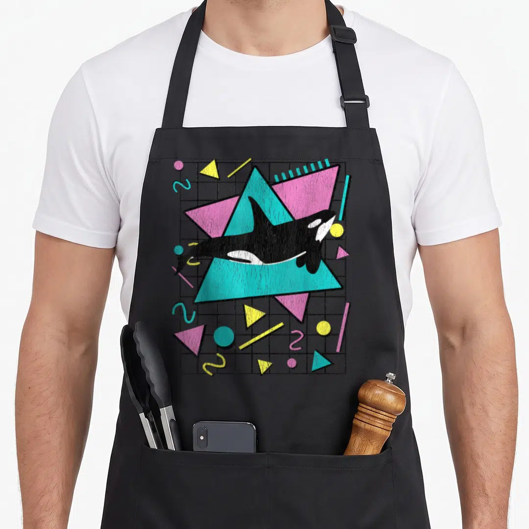 Orca 90s apron - Retro orca over teal and pink triangles on a black grid with scattered yellow shapes — shown on a black cotton canvas apron against a white background