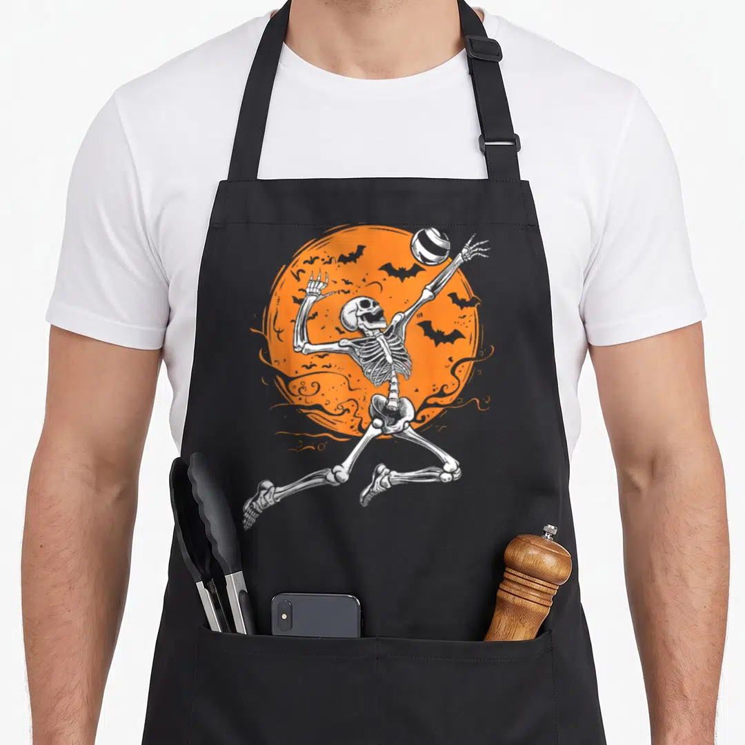 Volleyball skeleton halloween apron - White skeleton jumping to spike volleyball in front of orange moon with bats — shown on a black cotton canvas apron against a white background