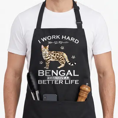 Bengal cat apron - Cartoon Bengal cat with I Work Hard So My Bengal Cat Can Have A Better Life text and paw prints — shown on a black cotton canvas apron against a white background
