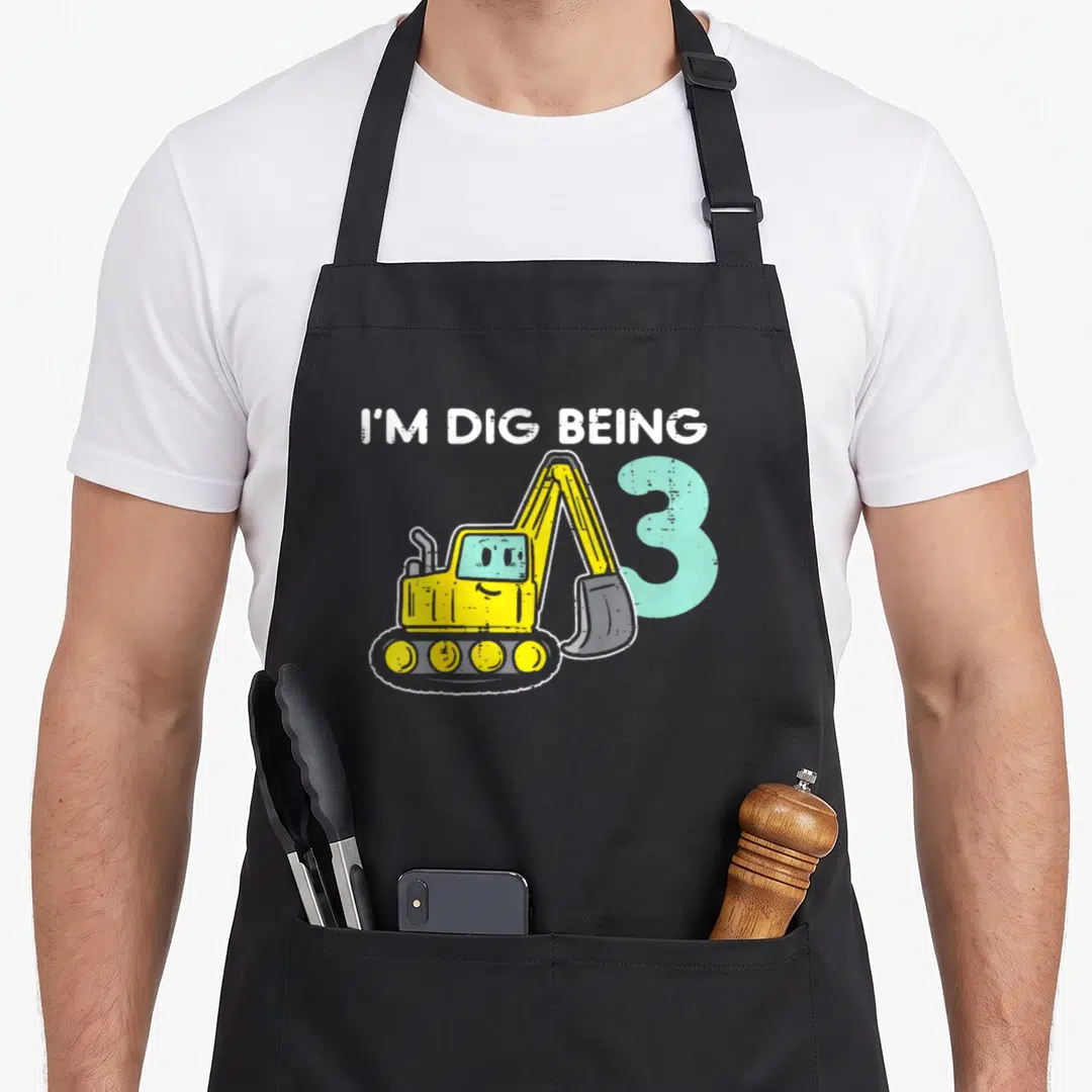 Dig birthday apron - Yellow cartoon excavator overlapping a light blue number 3 under arched white text reading I'M DIG — shown on a black cotton canvas apron against a white background