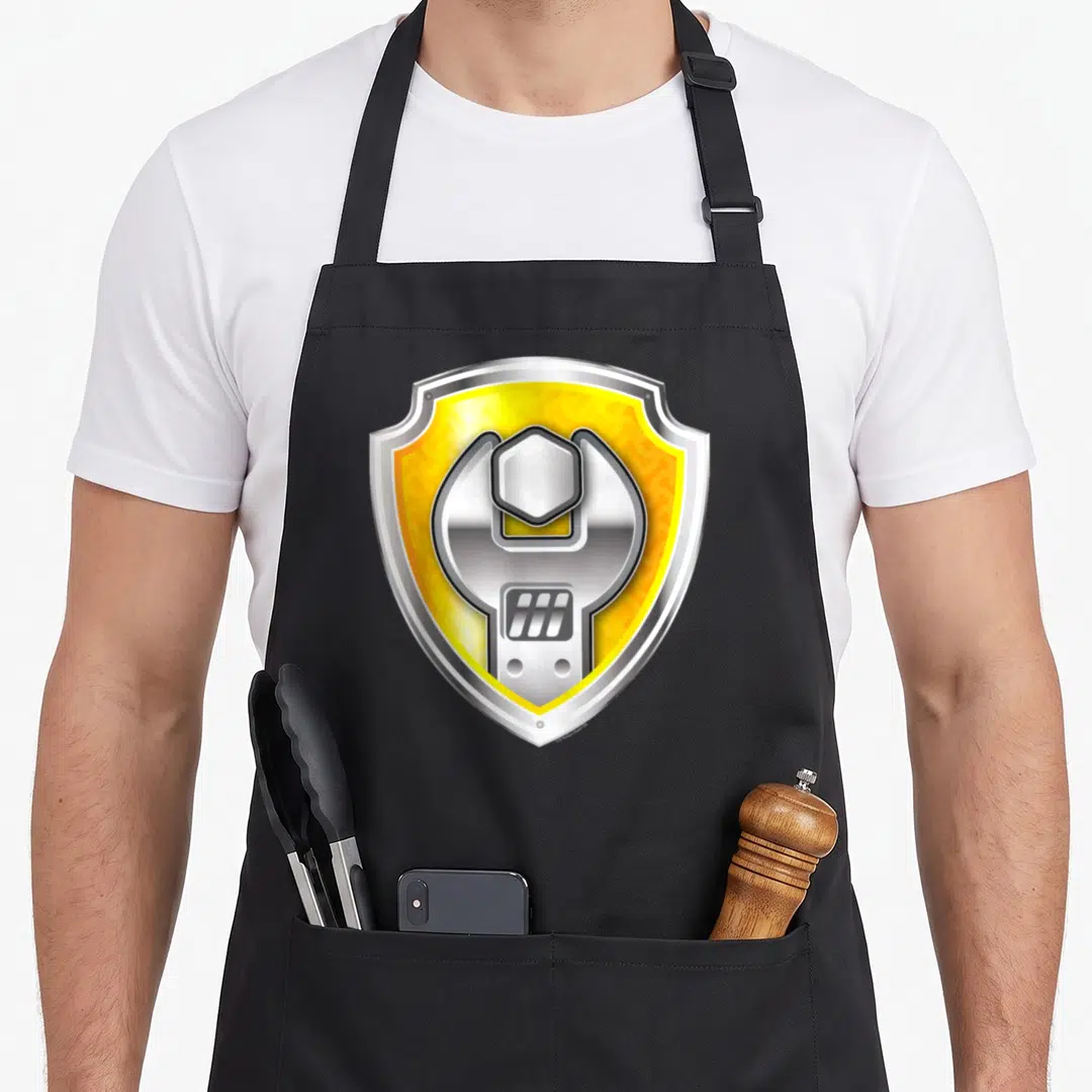 Rubble novelty apron - Silver wrench and nut inside a yellow shield with silver border — shown on a black cotton canvas apron against a white background