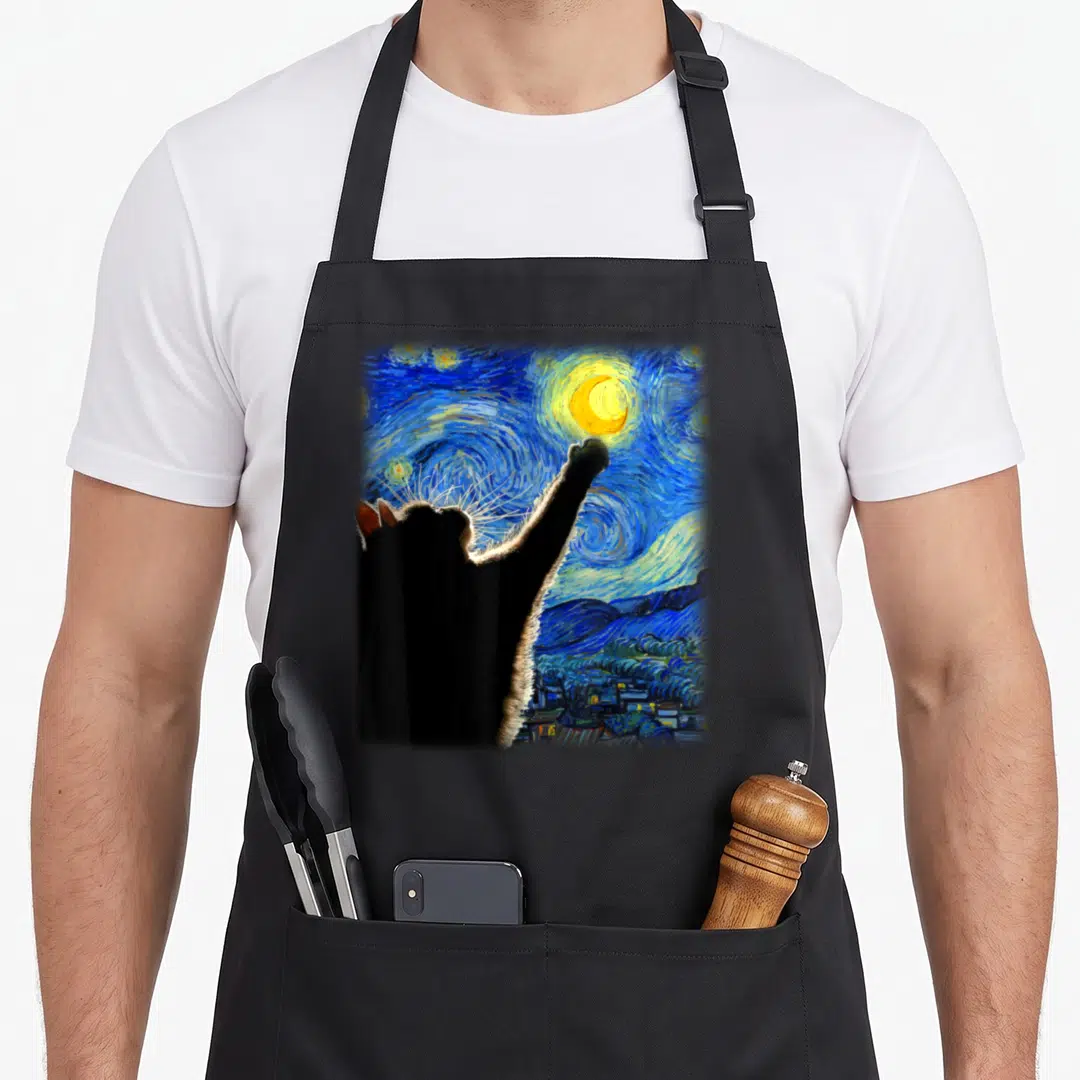 Starry cat in an apron - Black cat silhouette pawing at yellow crescent moon in swirling blue and yellow starry sky — shown on a black cotton canvas apron against a white background