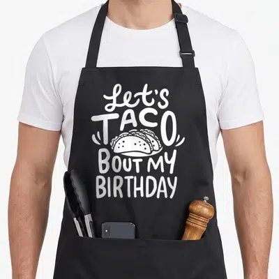 Birthday taco apron - White text Let's Taco Bout My Birthday with three tacos on plate — shown on a black cotton canvas apron against a white background