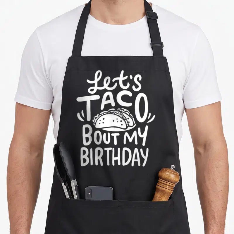 Birthday taco apron - White text Let's Taco Bout My Birthday with three tacos on plate — shown on a black cotton canvas apron against a white background