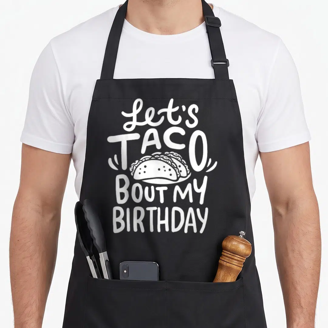 Birthday taco apron - White text Let's Taco Bout My Birthday with three tacos on plate — shown on a black cotton canvas apron against a white background