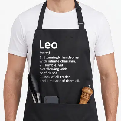 Leo definition birthday apron - White dictionary style text defining Leo as handsome, humble, and a master of trades — shown on a black cotton canvas apron against a white background