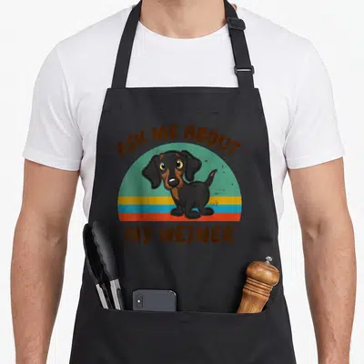 Ask dog apron - Cartoon dachshund on teal and orange stripes with brown text above and below — shown on a black cotton canvas apron against a white background
