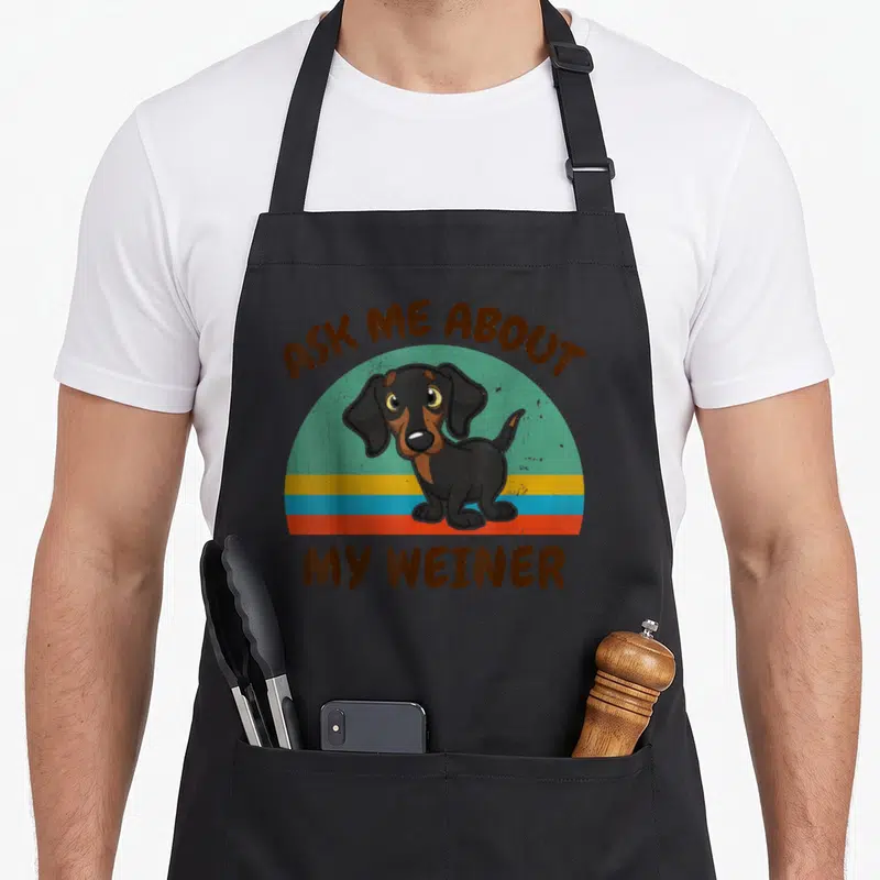 Ask dog apron - Cartoon dachshund on teal and orange stripes with brown text above and below — shown on a black cotton canvas apron against a white background