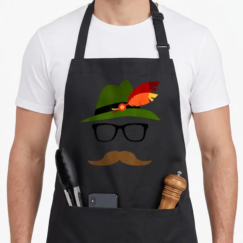 German oktoberfest lederhosen apron - Minimalist face with green hat, red feather, black glasses, brown mustache — shown on a black cotton canvas apron against a white background