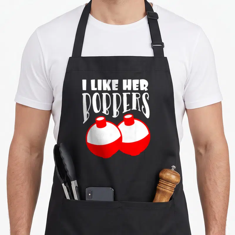 Bobber couple apron - I LIKE HER BOBBERS text above two red and white cartoon fishing floats — shown on a black cotton canvas apron against a white background