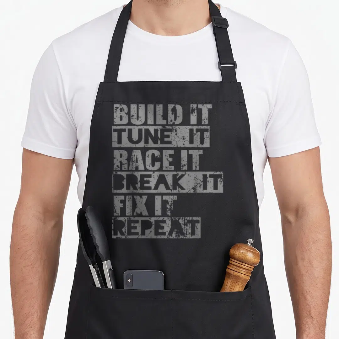 Build air show apron - Stacked grey text reading BUILD IT TUNE IT RACE IT BREAK IT FIX IT REPEAT on black fabric — shown on a black cotton canvas apron against a white background